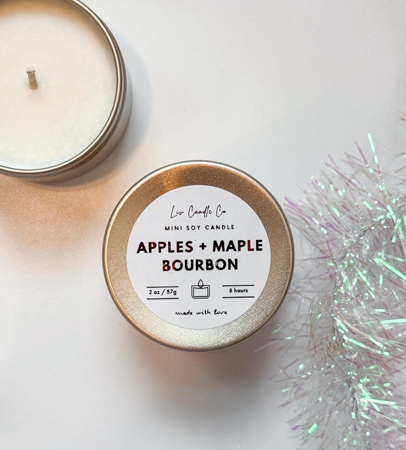 Puede incluir: Una lata de vela de soja dorada en miniatura con una etiqueta blanca que dice "Liv Candle Co. MINI SOY CANDLE APPLES + MAPLE BOURBON 2 oz/57g 8 hours made with love".