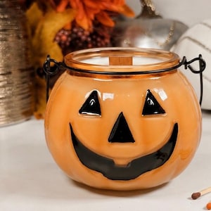 May include: Orange pumpkin-shaped candle holder with a black smiling face. The candle holder has a glass lid and a black metal handle. Matches are scattered in front of the candle holder. Other decorative items are in the background.