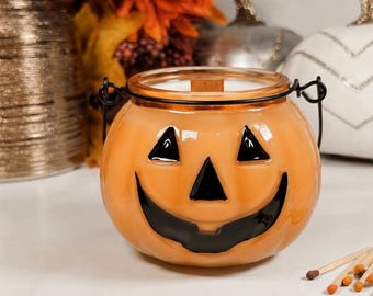 May include: Orange pumpkin-shaped candle holder with a black smiling face. The candle holder has a glass lid and a black metal handle. Matches are scattered in front of the candle holder. Other decorative items are in the background.