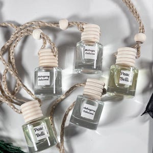 May include: Five small, clear glass bottles with wooden caps and labels, suspended by a beige rope. The labels read "Mahogany Oakwood" and "Peach Bellini." The bottles are filled with a light-coloured liquid. The background is white.