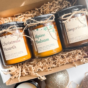 May include: A gift box containing three glass jar candles with black lids. The labels read "Christmas Hearth", "Alpine Balsam", and "Gingerbread". Each candle is tied with twine. The candles are filled with a yellow wax and are 221 g.