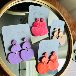 May include: A collection of handmade clay earrings in various colors, including coral, lavender, and peach. Each pair features a unique three-tiered design with gold-colored metal findings. The earrings are displayed on gray cards with the "LA BODEGA COMPANY" logo.