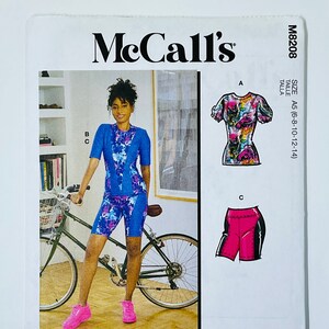 May include: McCall's sewing pattern M8208 featuring a woman in a blue and purple patterned top and shorts, standing next to a bicycle. The pattern includes multiple view options with simple instructions. The size is A5 (6-8-10-12-14).