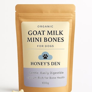 Op de afbeelding: Een bruine papieren zak met hondensnacks, met het opschrift "Organic Goat Milk Mini Bones" met het Honey's Den-logo. De zak heeft een crème label met blauwe accenten en bevat de tekst "Gentle, Easily Digestible, Calcium-Rich for Bone Health, 100g". Verschillende botvormige traktaties liggen verspreid.