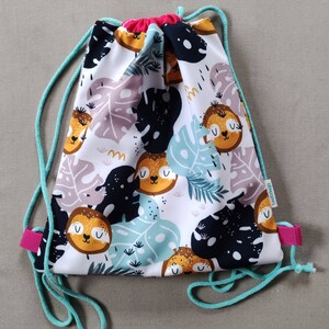 Può includere: Borsa a sacco bianca con una stampa colorata di bradipi e foglie tropicali. La borsa ha laccetti di colore turchese e dettagli rosa.