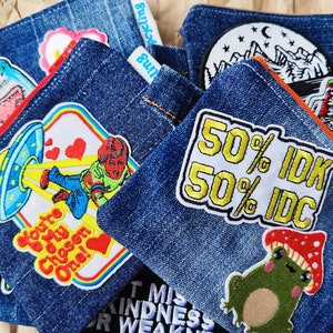 May include: A set of six denim pouches with various embroidered patches. The patches feature a shark, a UFO, a person riding a skateboard, a frog, a mushroom, and text that says "50% IDK 50% IDC".