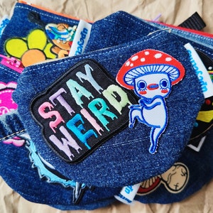 May include: A blue denim pouch with a variety of colourful patches, including a mushroom character, a flower, and a patch that says "Stay Weird".