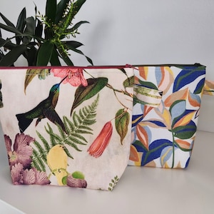 Könnte beinhalten: Zwei Reißverschlusstaschen mit Blumenmustern. Eine Tasche zeigt einen Kolibri und tropische Pflanzen auf cremefarbenem Hintergrund. Die andere Tasche hat ein blaues, grünes und orangefarbenes Blattmuster auf weißem Hintergrund. Beide Taschen haben einen roten Reißverschluss.