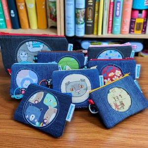 May include: A collection of denim zipper pouches with various circular fabric appliqués. Designs include a cat, bears, a mushroom, and floral patterns. The pouches are arranged on a wooden surface, with a bookshelf in the background.