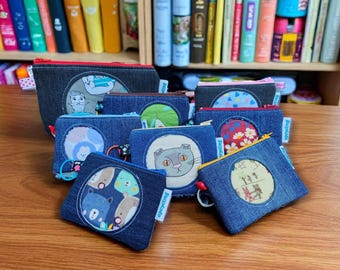 May include: A collection of denim zipper pouches with various circular fabric appliqués. Designs include a cat, bears, a mushroom, and floral patterns. The pouches are arranged on a wooden surface, with a bookshelf in the background.