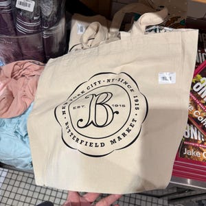 Puede incluir: Un bolso tote de color crema con texto negro que dice "New York City NY Since 1915 Est. Butterfield Market". El bolso tiene dos asas y está hecho de un material de lona resistente. Otros artículos son visibles en el fondo.