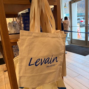 May include: Cream-colored tote bag with long handles, featuring the blue text "Levain BAKERY". The bag is hanging on a wooden display shelf, with other bags and packaged goods visible. The background includes a storefront.