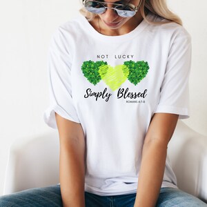 Op de afbeelding: Wit T-shirt met een ontwerp van een groene klaver en de tekst "Not Lucky Simply Blessed Romans 4:7-8"