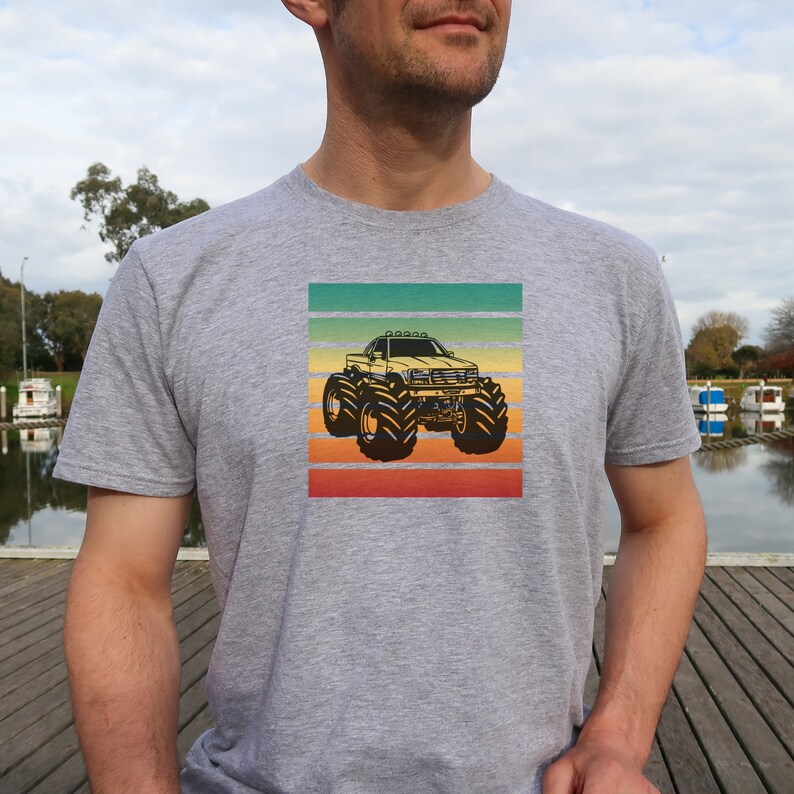 May include: A gray t-shirt with a retro-style graphic of a monster truck with large tires. The graphic is set against a background of horizontal stripes in shades of green, yellow, orange, and red.