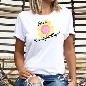 Peut inclure: T-shirt blanc avec un motif de fleur jaune et le texte "It's a Beautiful Day !". La fleur a un centre rose qui ressemble à une balle de pickleball.