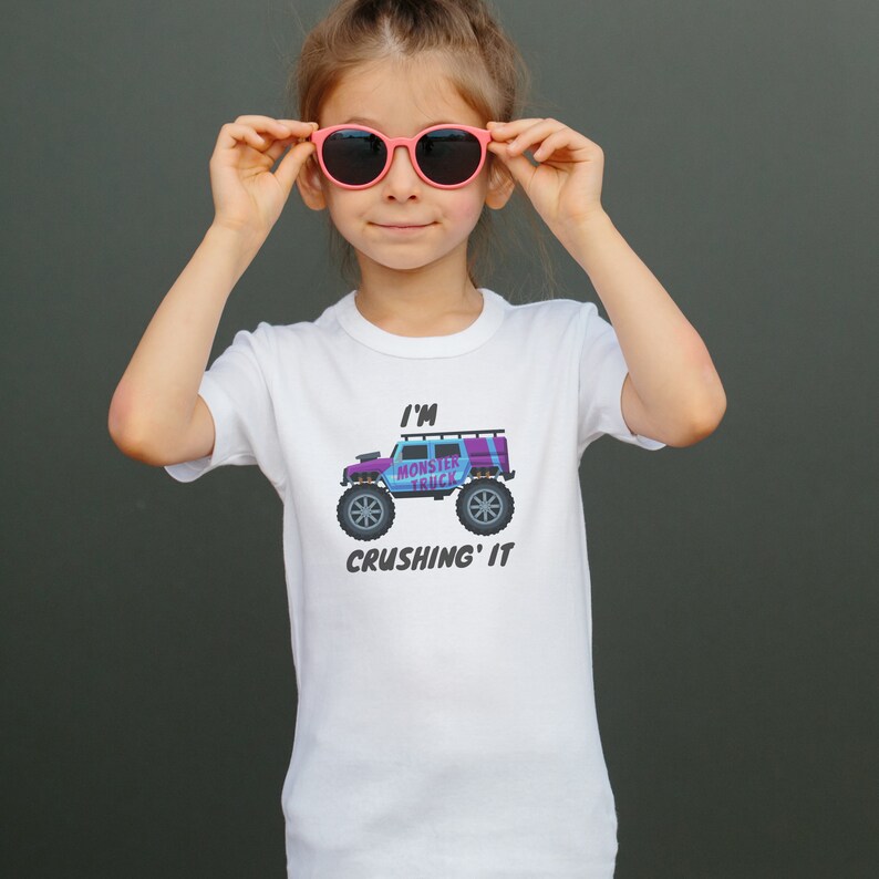 May include: A white t-shirt featuring a colorful monster truck graphic with the text "I'M MONSTER TRUCK CRUSHING IT." The shirt is worn by a person wearing pink sunglasses.