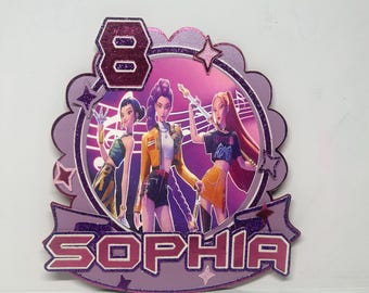 May include: A purple and pink cake topper featuring three animated figures, the number 8, and the name "SOPHIA". The design includes a glittery border and star accents, with a musical note background.