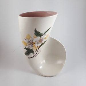 May include: A white ceramic vase with a pink rim and a floral design. The vase is shaped like a curved tube with a wide opening at the top and a narrow opening at the bottom. The floral design features white flowers with yellow centres and green leaves.