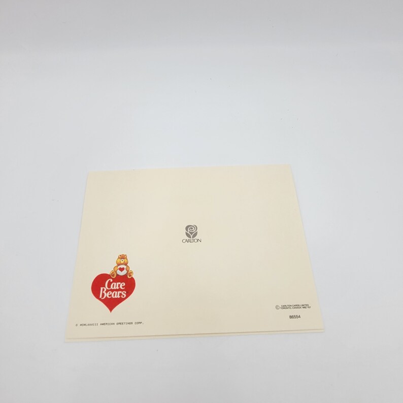 May include: An off-white greeting card with the Care Bears logo, a red heart, and a bear illustration. The Carlton logo and the number 86554 are also printed on the card. The card is folded.