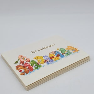 May include: A stack of greeting cards with a cream background. The cards feature a colourful illustration of cartoon bears pulling a sled, with the text "It's Christmas!" printed on the front.