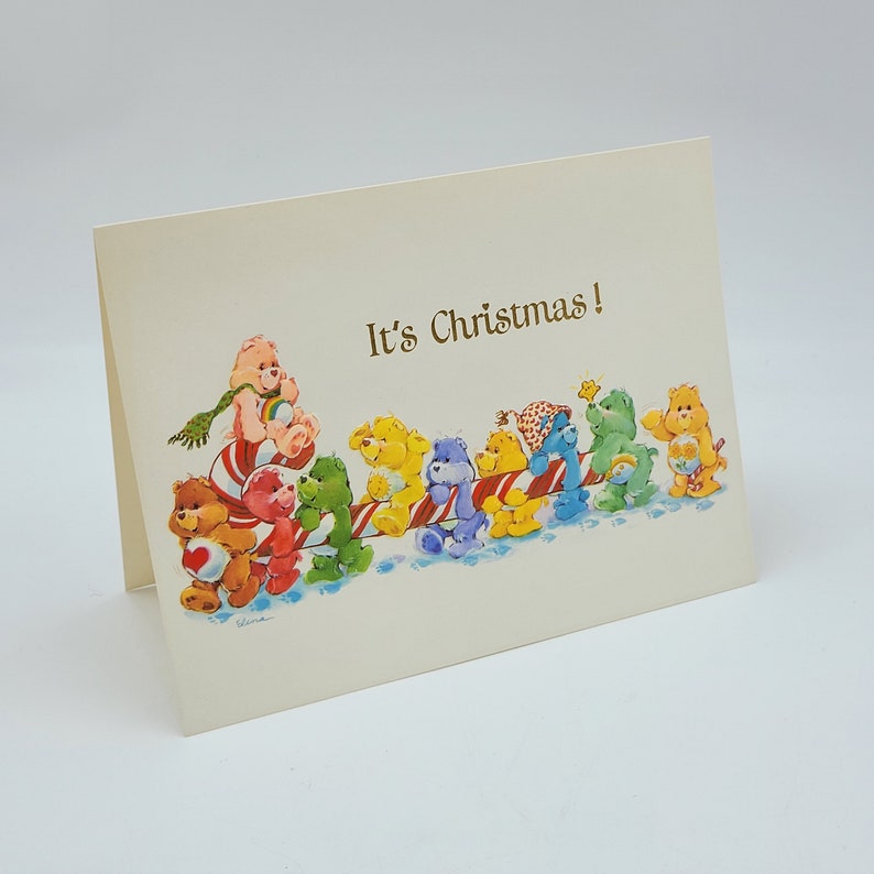 May include: Cream Christmas card with a line of colourful Care Bears on a candy cane sled. The gold text reads "It's Christmas!". The bears are in various colours, including pink, yellow, blue, and green, creating a festive scene.