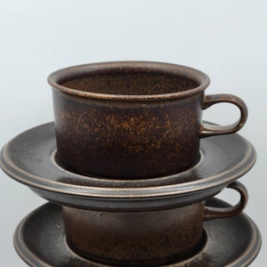 Peut inclure: Deux tasses à café en céramique marron empilées avec des soucoupes assorties. Les tasses ont une forme cylindrique avec une petite anse. Les soucoupes sont rondes avec un bord légèrement surélevé. Les tasses et les soucoupes ont un aspect tacheté et texturé.