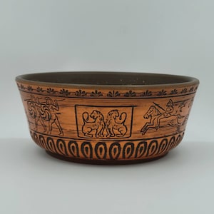 May include: A terracotta bowl with a brown glaze and black painted designs. The bowl features a repeating pattern of stylised lions, horses, and warriors.