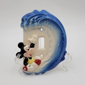 May include: A ceramic light switch cover featuring Mickey Mouse riding a surfboard in a blue wave. The switch cover is white with a blue wave design and Mickey Mouse is wearing a yellow life vest.