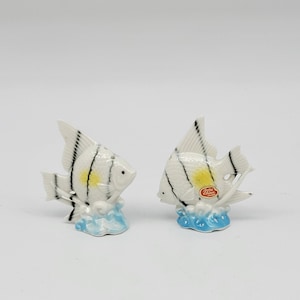 May include: A pair of white ceramic salt and pepper shakers shaped like angelfish with black stripes and yellow accents. The shakers are sitting on a blue and white wave base. The base of the right shaker has a red label that says "Bone China Japan".