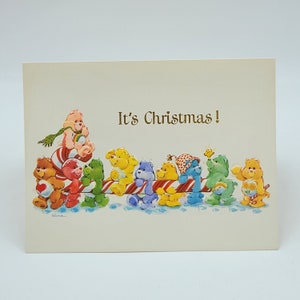 May include: A vintage Christmas card with the text "It's Christmas!" features a line of colourful Care Bears. The bears are arranged on a candy cane-striped rail, with a light beige background. The bears are in various colours and poses.