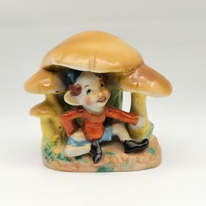 May include: A ceramic figurine of a small elf-like figure under a mushroom. The elf wears an orange shirt, blue shorts, and black shoes. The mushroom has a tan cap and a yellow stem, with green and brown accents.