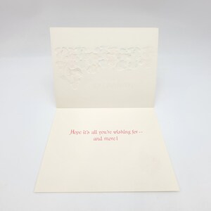 May include: A cream-coloured greeting card, open to reveal red text: "Hope it's all you're wishing for -- and more!" The top section features an embossed floral design. The card is set against a plain white background.
