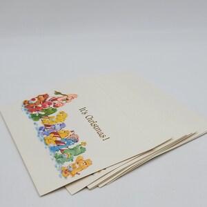 May include: A pile of cream-coloured Christmas cards. The cards feature a colourful illustration of cartoon bears and the text "It's Christmas!" in gold lettering. The cards are stacked on a white surface.