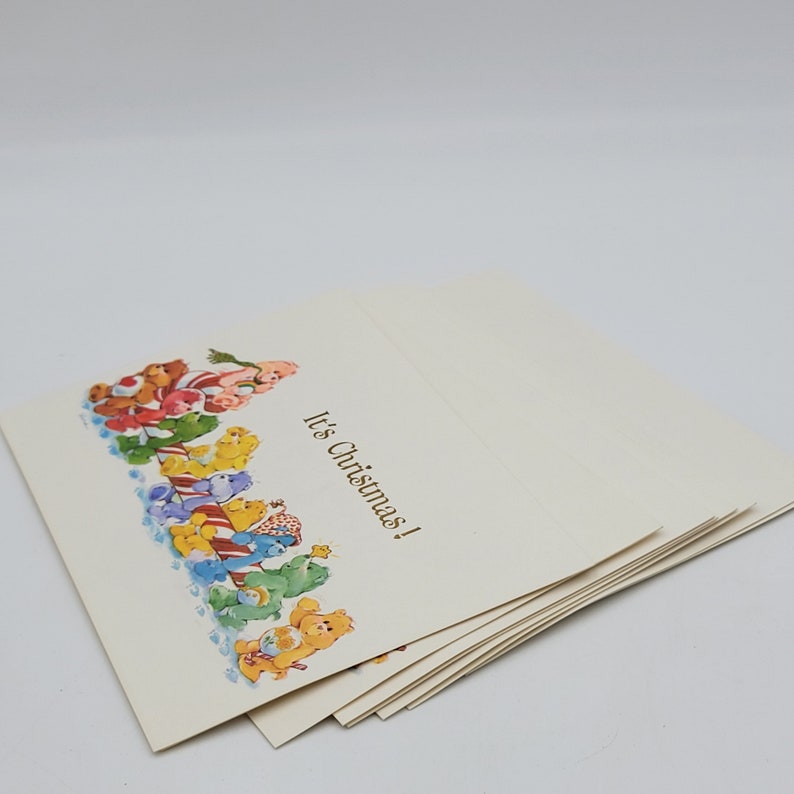 May include: A pile of cream-coloured Christmas cards. The cards feature a colourful illustration of cartoon bears and the text "It's Christmas!" in gold lettering. The cards are stacked on a white surface.