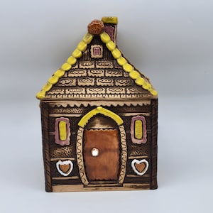 May include: A ceramic gingerbread house cookie jar with a brown, yellow and pink glaze. The roof is decorated with a yellow icing design and the sides have a brown woodgrain pattern. The house has a door, two windows and a chimney.