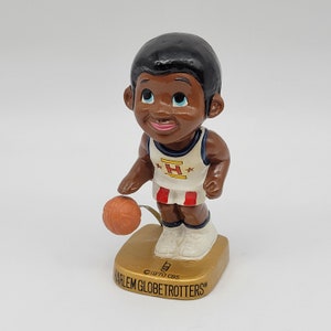 Peut inclure: Une figurine de joueur de basket-ball des Harlem Globetrotters à tête oscillante. Le joueur porte un maillot blanc avec des bordures rouges et bleues, un bandeau rouge et bleu et un short noir. Le joueur tient un ballon de basket dans sa main droite et sourit. Le socle du bobblehead est doré et porte les inscriptions "Harlem Globetrotters" et "©1970 CBS".