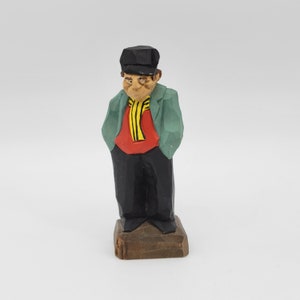 May include: A hand-carved wooden figurine of a person wearing a black hat, green jacket, red shirt, and yellow and black scarf. The figurine is standing on a wooden base.