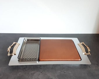 May include: A silver serving tray with two compartments. The left compartment has a clear glass insert with a diamond pattern. The right compartment has a brown leather insert. The tray has two gold handles.