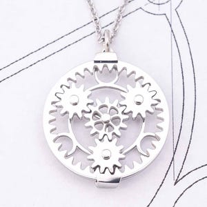 May include: A silver-colored pendant necklace featuring a circular design of interlocking gears. The pendant has a detailed, mechanical aesthetic, with multiple gears of varying sizes. The necklace is displayed on a white surface with faint blueprint-like markings.