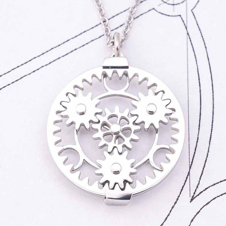 May include: A silver-colored pendant necklace featuring a circular design of interlocking gears. The pendant has a detailed, mechanical aesthetic, with multiple gears of varying sizes. The necklace is displayed on a white surface with faint blueprint-like markings.