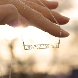 Puede incluir: Un collar de cadena de plata con un colgante de barra de oro con el número "299.792.458 m/s" grabado en él.