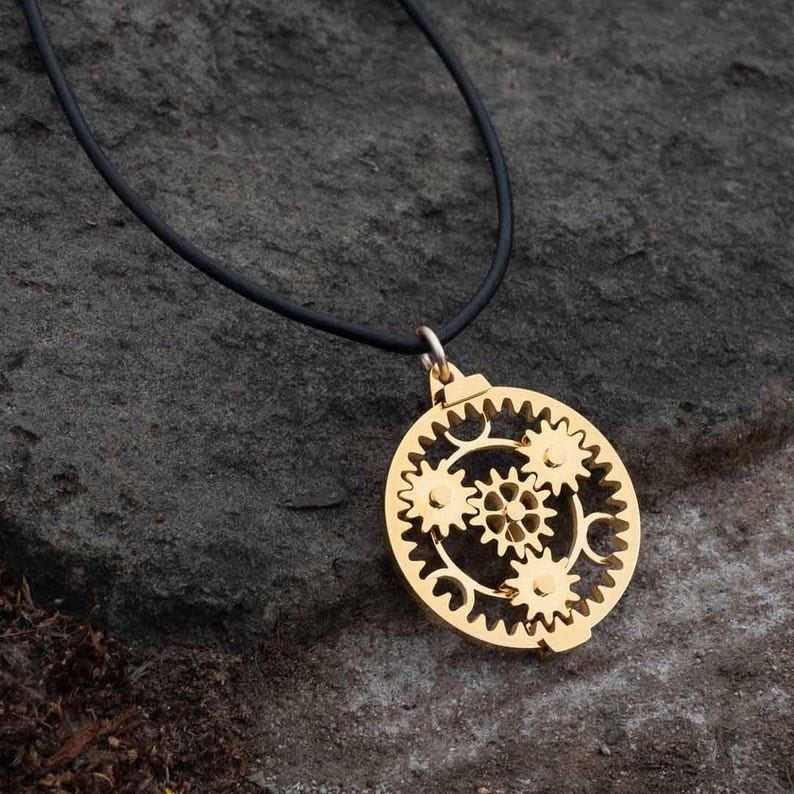 May include: A gold-colored pendant necklace featuring a circular design of interlocking gears. The pendant hangs from a black cord. The necklace is displayed on a textured, dark gray surface.