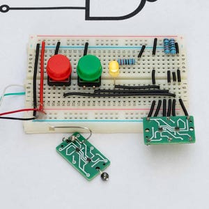 Op de afbeelding: Een elektronica project op een wit breadboard met rode en groene drukknoppen, een gele LED en diverse elektronische componenten. Twee groene printplaten en een printplaat sleutelhanger zijn ook aanwezig.