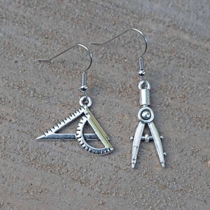 May include: A pair of silver earrings featuring a protractor and compass design. The earrings are dangle style with a silver hook.