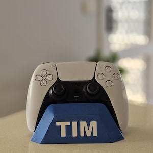 May include: A white and black PlayStation 5 controller on a blue stand. The stand displays the name "TIM" in white. The controller features a modern design with button symbols.