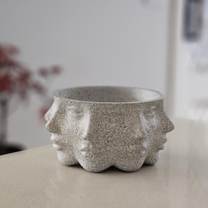 Peut inclure: Un bol décoratif gris moucheté avec trois visages sculptés à l'extérieur. Les visages ont les yeux fermés et une expression sereine. Le bol est posé sur une surface de couleur claire.