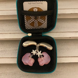 Peut inclure: Une bo&icirc;te de transport turquoise avec un int&eacute;rieur noir. La bo&icirc;te contient deux aides auditives, deux embouts auriculaires roses avec des breloques d'&eacute;toiles de mer argent&eacute;es scintillantes et une petite carte blanche sur laquelle est inscrit "high level hearing".