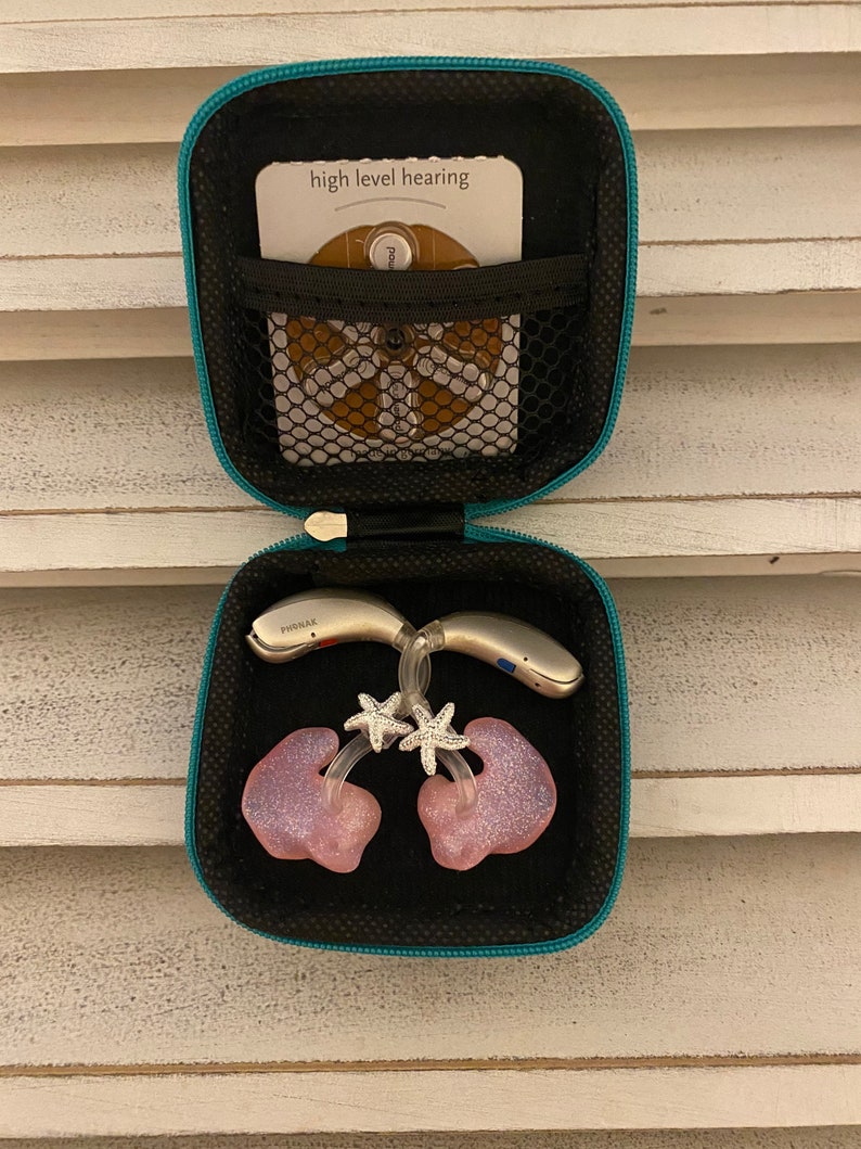 Peut inclure: Une bo&icirc;te de transport turquoise avec un int&eacute;rieur noir. La bo&icirc;te contient deux aides auditives, deux embouts auriculaires roses avec des breloques d'&eacute;toiles de mer argent&eacute;es scintillantes et une petite carte blanche sur laquelle est inscrit "high level hearing".