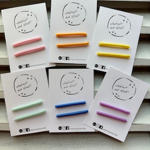 Könnte beinhalten: Sechs Karten mit dem Text "whatnot and Wonder" und einem Paar Perlenhaarspangen in verschiedenen Farben: rosa, orange, gelb, hellgrün, blau und lila.