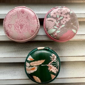 May include: Three round, zippered cases with floral and feather designs. The top case is light pink with a peony pattern. The middle case has a brown background with pink flowers. The bottom case is dark green with feather and floral accents.